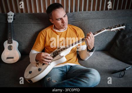 Man playing guitar while sitting on sofa, musical hobby and relaxed home atmosphere. Stock Photo