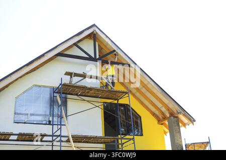 Worker on multi-level scaffold installs facade trim, yellow and white house, crafting modern exterior, teamwork and renovation project concept Stock Photo