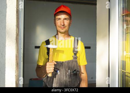 Portrait of construction worker in red cap holding hammer, ready for carpentry, nailing, and home repairs, handyman business, tool kit on-site Stock Photo