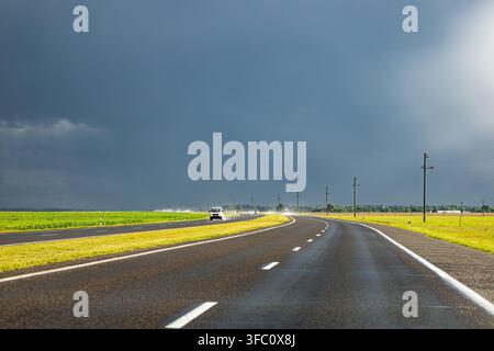 A smooth asphalt highway curves through open farmland under a massive dark storm front. Sunlit green and golden fields contrast with the deep blue sky Stock Photo