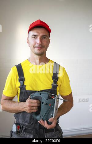Repairman in red cap holding cordless drill, house facade, versatile tools for repairs, handyman services, at-home work, building maintenance and inst Stock Photo