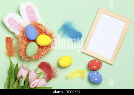Easter eggs in a nest, bunny ears, flowers and a frame with copy space on a mint green background Stock Photo