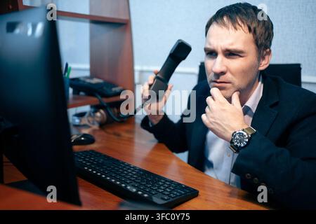 Businessman holding a landline phone receiver, looking thoughtfully into the distance with his hand on his chin. Business call, deep thinking, and pro Stock Photo