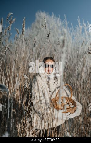 Woman in white coat stands in tall grass holding a basket under a clear blue sky in winter Stock Photo