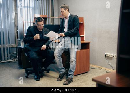 Two businessmen reviewing a document together in an office, one standing and one sitting. Teamwork, professional discussion, and corporate analysis. Stock Photo