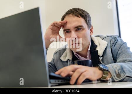 Focused man working on a laptop, concentrating on online tasks in a modern office. Business, remote work, productivity and problem solving concept. Stock Photo
