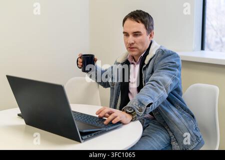 Businessman working remotely on a laptop in a modern cafe, focused on online tasks in a comfortable coworking environment with natural daylight. Stock Photo
