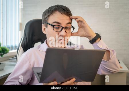 Confused office worker touching his forehead while looking at his laptop screen with a skeptical expression. Misunderstanding, difficult task, or read Stock Photo
