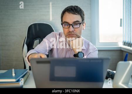 Focused businessman sitting at his desk with his chin resting on his fist, looking intently at his laptop screen. Deep concentration, online business Stock Photo