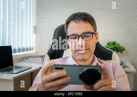 Smiling man wearing glasses using smartphone in landscape orientation while relaxing in office, enjoying mobile entertainment. Stock Photo