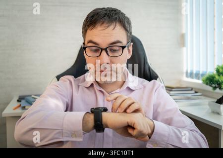 Man in a pink shirt and glasses checking the time on his smartwatch while sitting at an office desk. Stock Photo