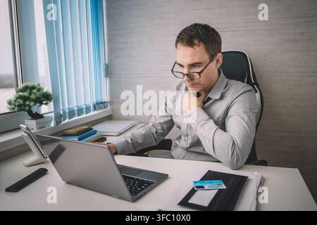 Thoughtful businessman stroking his beard while looking at his laptop screen. Deep analysis, decision making, and professional reflection at work. Stock Photo