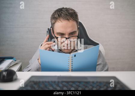 Focused worker talking on smartphone while reviewing notes in office, managing business tasks, planning, and communication during busy workday. Stock Photo