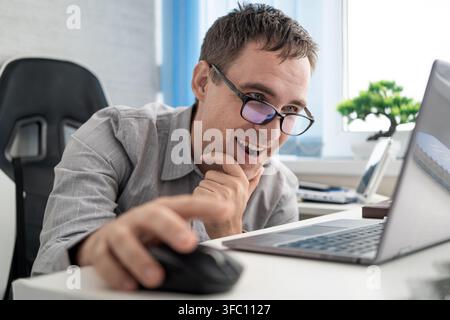 Happy worker smiling while using a laptop and computer mouse in office, enjoying successful online work, positive results, and productive digital comm Stock Photo