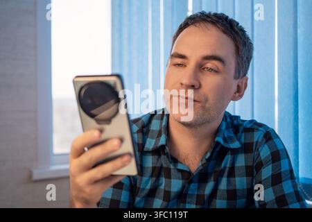 Young adult man wearing blue plaid shirt looking at smartphone held in right hand while seated near window with vertical blinds, soft natural daylight Stock Photo