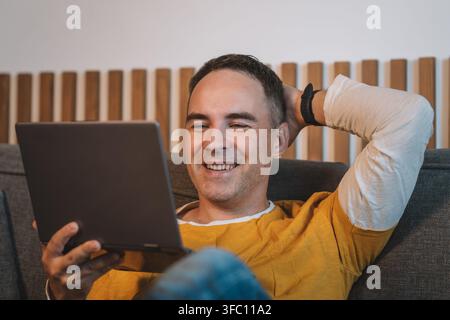 Middle-aged man reclining on gray sofa smiling while holding tablet at arm's length, relaxed casual pose conveying contentment and engagement, wearing Stock Photo