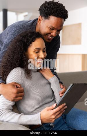 Couple sitting on gray sofa, wearing gray sweater and dark knit, holding black tablet, smiling. Home, livingroom, minimalist, modern, neutral, cozy, d Stock Photo