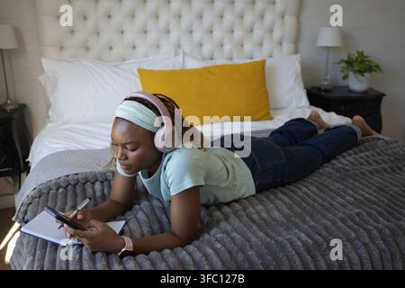 Female lying on bed wearing pink headphones, scrolling smartphone and writing in notebook with pen Stock Photo