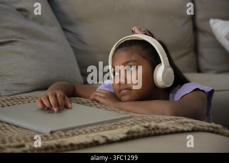 African American youth female lying on living room couch, wearing headphones, touching laptop lid Stock Photo