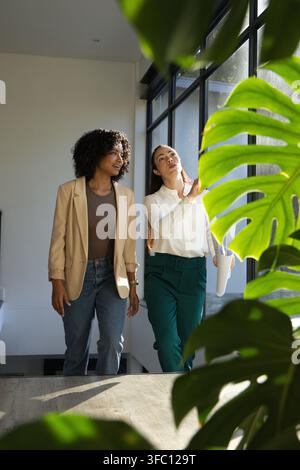Diverse female coworkers walking and discussing while holding rolled plans in workwear by window Stock Photo