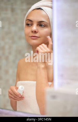 Young woman looking into mirror, wearing a towel and applying moisturizer to her face. Self-care and beauty routine Stock Photo