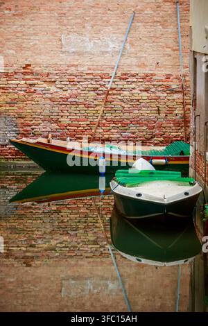 Two small boats rest on calm canal water beside an aged brick wall in Venice, Italy, with clear reflections and a quiet historic mood. Stock Photo