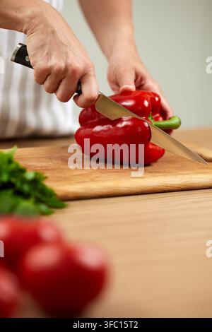 Female hands slicing fresh red bell pepper with knife on a cutting board, preparing healthy meal in a home kitchen Stock Photo