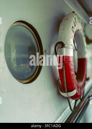 Close-up detail of a ship's side featuring a weathered porthole and a red and white lifebuoy with rope, hanging on the vessel's exterior in Gdynia, Po Stock Photo