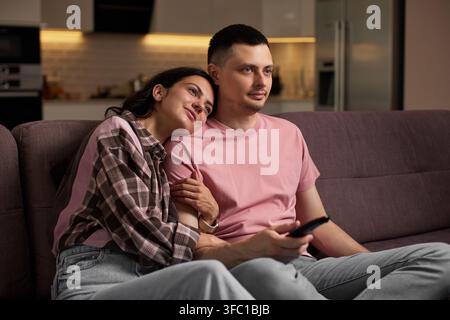 Young couple enjoying leisure time on sofa, watching television and embracing in comfortable living room Stock Photo