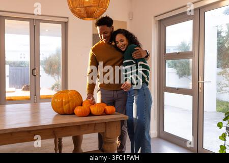 African American couple standing embracing in dining area beside table with pumpkins, pendant light. Affection, partners, cozy, autumn, gourds, plants Stock Photo