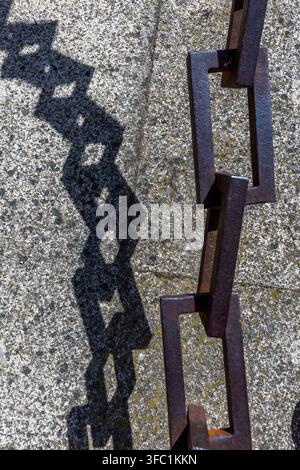 A heavy iron chain casts a sharp, geometric shadow onto a textured stone surface outdoors in Spain. Stock Photo