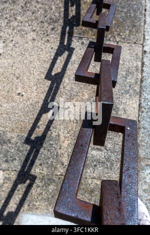 A heavy iron chain casts a sharp, geometric shadow onto a textured stone surface outdoors in Spain. Stock Photo