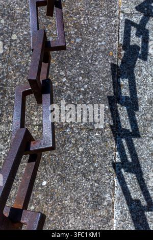 A heavy iron chain casts a sharp, geometric shadow onto a textured stone surface outdoors in Spain. Stock Photo