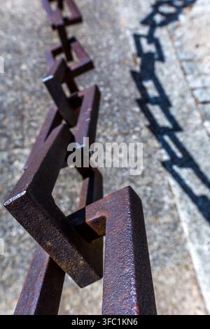 A heavy iron chain casts a sharp, geometric shadow onto a textured stone surface outdoors in Spain. Stock Photo