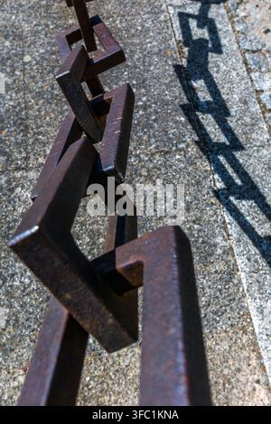 A heavy iron chain casts a sharp, geometric shadow onto a textured stone surface outdoors in Spain. Stock Photo