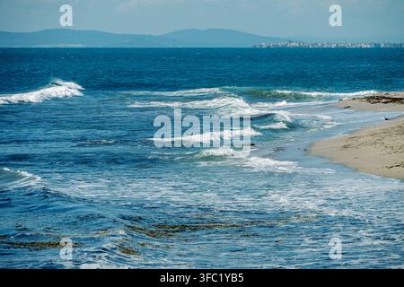Gentle blue ocean waves roll onto a sandy shoreline, with a distant coastal city and hazy mountains visible on the horizon under a clear sky. Stock Photo