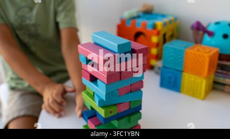 Colorful wooden blocks stacked into a tall tower on a table while a child plays with toys in the background at home. Stock Photo