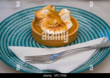 a freshly baked mini lemon meringue tart garnished with candied peel sits on a blue patterned ceramic plate alongside a white napkin and a silver dess Stock Photo