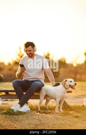 Happy man relaxing in park with dog, enjoying music during golden hour Stock Photo