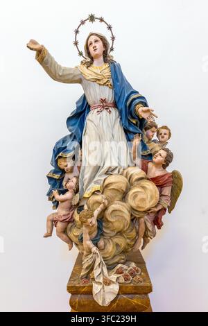 An ornate wooden sculpture of the Virgin Mary surrounded by cherubs and clouds stands against a white background in Spain. Stock Photo