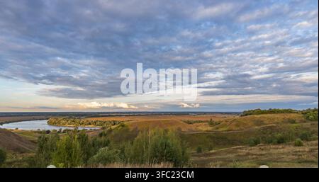 Colorful landscape in the evening, gorgeous blue sky with golden clouds over a field with hills and a river. A magical moment of nature. Stock Photo