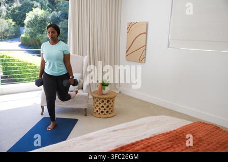 African American woman doing split squat on blue mat in bedroom using armchair holding hex weights Stock Photo