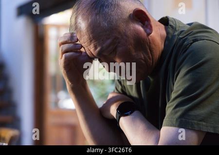 Senior Asian man leaning on right hand, wearing olive-green T-shirt and black smartwatch at home Stock Photo