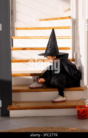 African child sitting on stairs tying sneaker in pointed witch hat with jack o lantern nearby Stock Photo