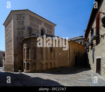 Stone church with arched windows and textured walls. Cobbled street curves around ancient structure. Sunlight casts long shadows on historic pavement. Stock Photo