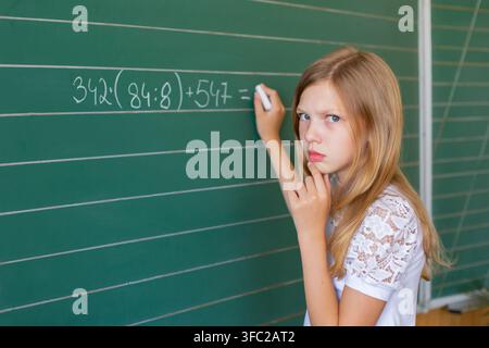 Girl in a white shirt gestures with one finger while speaking in front of a green school board. She seems engaged in learning. Stock Photo
