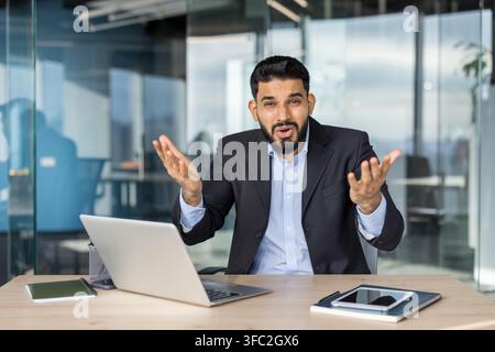 Businessman sitting at an office desk, looking directly at the viewer with a confused expression, shrugging with open hands, showing uncertainty or disbelief while working on a laptop Stock Photo