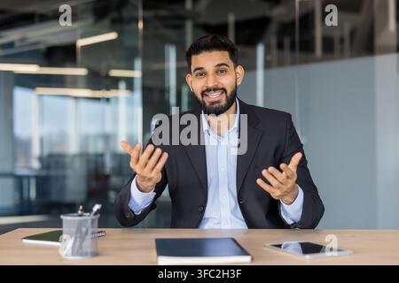 Young indian businessman discussing ideas, gesturing with hands while communicating virtually, explaining business strategy during an online meeting from a modern office workspace Stock Photo