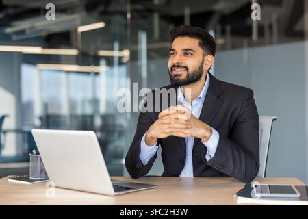 Young indian businessman sitting at office desk with laptop, smiling and looking away, contemplating strategies, leadership, and professional growth in a modern corporate environment Stock Photo