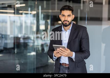 Young businessman standing confidently in a contemporary office setting, holding a digital tablet and looking directly at the viewer, representing corporate leadership and technology use Stock Photo
